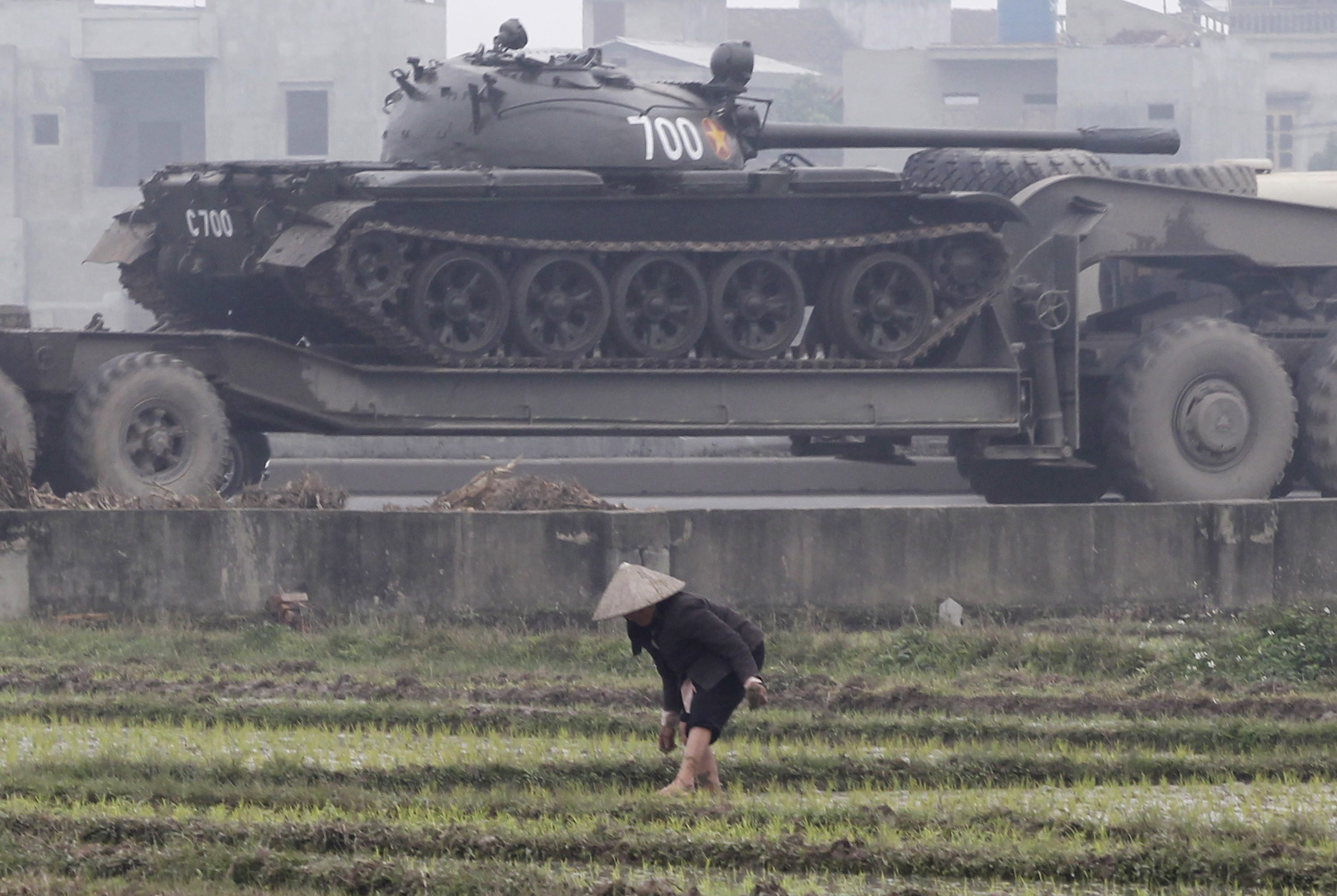 A military truck carries a tank past a farmer.