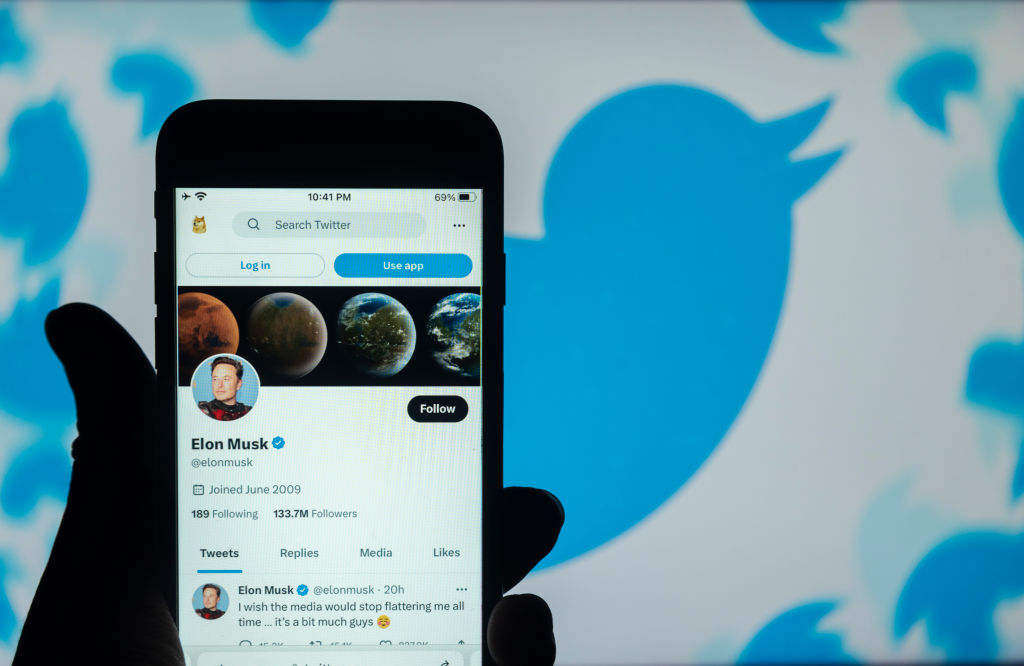 Elon Musk&#039;s twitter account is seen displayed on a mobile phone screen with a twitter logo in the background.